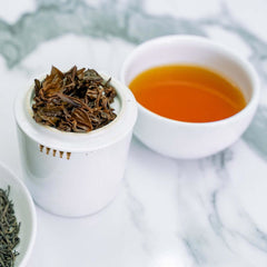 A white ceramic tea cup filled with amber-colored tea sits beside a small white container holding dried loose-leaf tea, with more tea leaves visible in a partial bowl on a light marble surface.